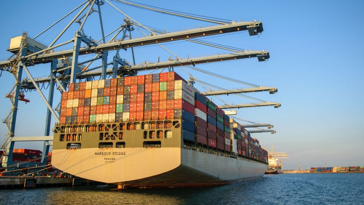 A large container ship at an industrial harbor with cranes under a clear blue sky.