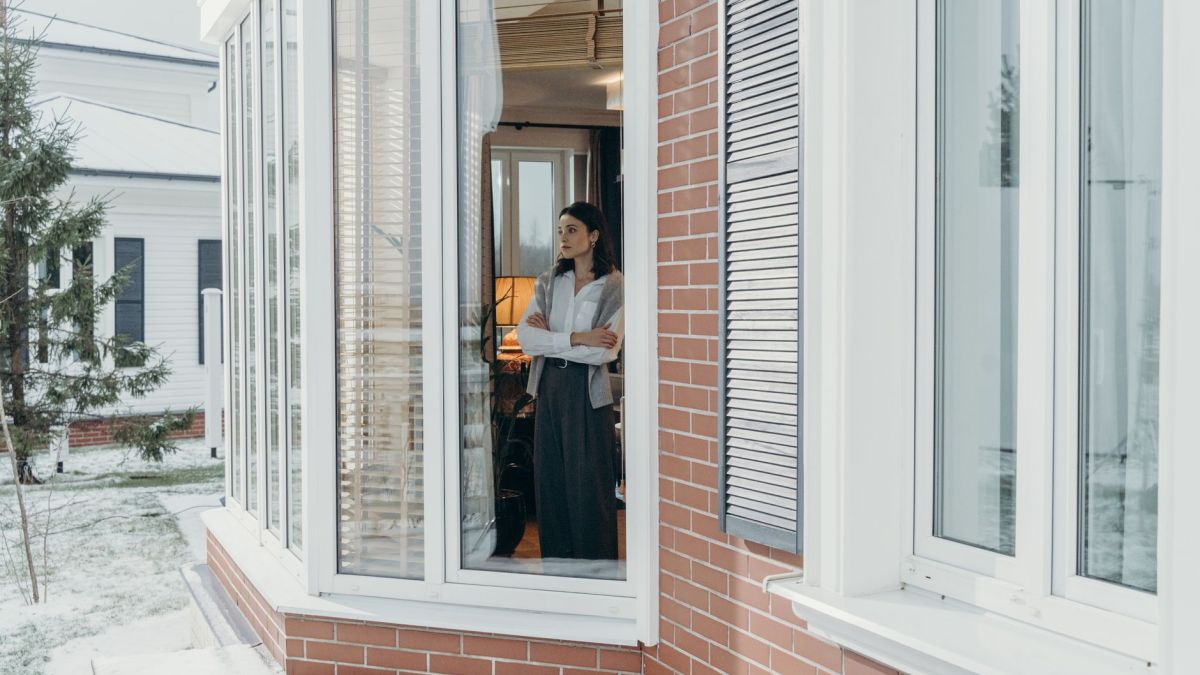 A woman stands indoors, arms crossed, looking out large windows on a snowy day, reflecting indoors and outdoors contrast.