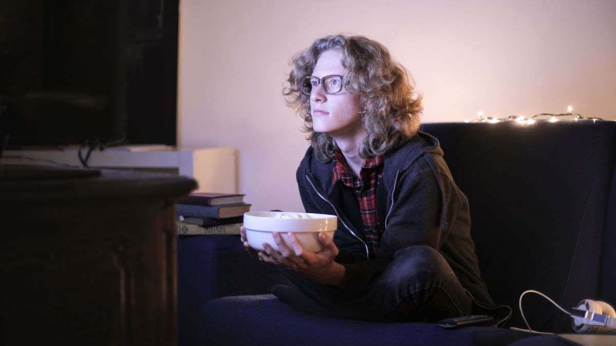 Serious young male with long curly hair sitting with crossed legs on couch in living room and enjoying interesting movie while chilling at home during weekend