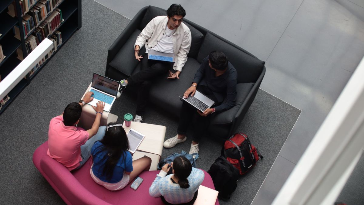 A group of diverse students using laptops and books in a library setting, studying together.