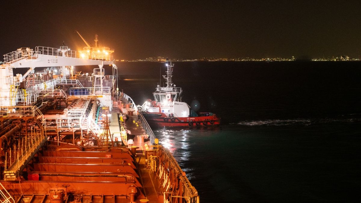 A vivid night scene of a cargo ship being assisted by a tugboat on the calm sea, illuminated by city lights.