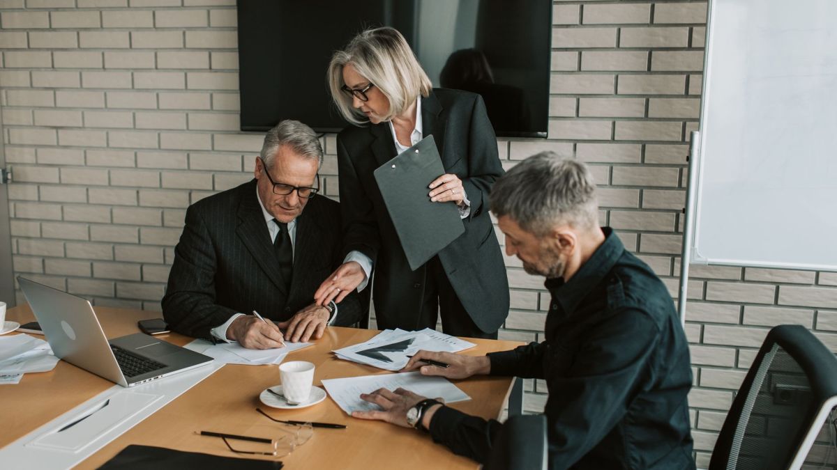 Three mature professionals in a business meeting discussing and signing documents in an office setting.