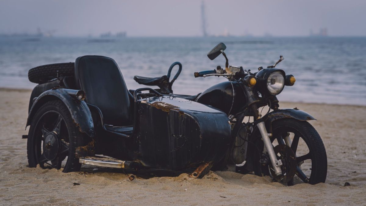 Rusty vintage motorcycle with sidecar parked on a sandy beach by the sea.