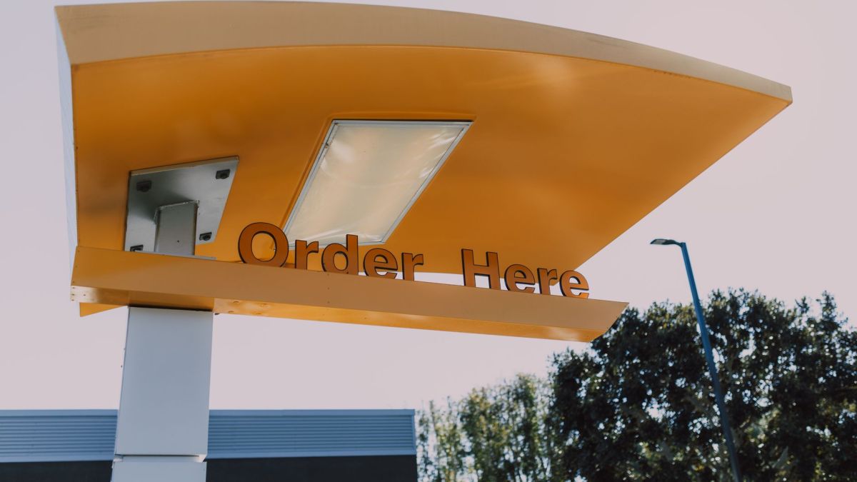 A modern drive-thru order sign with yellow canopy under a clear sky, surrounded by trees.