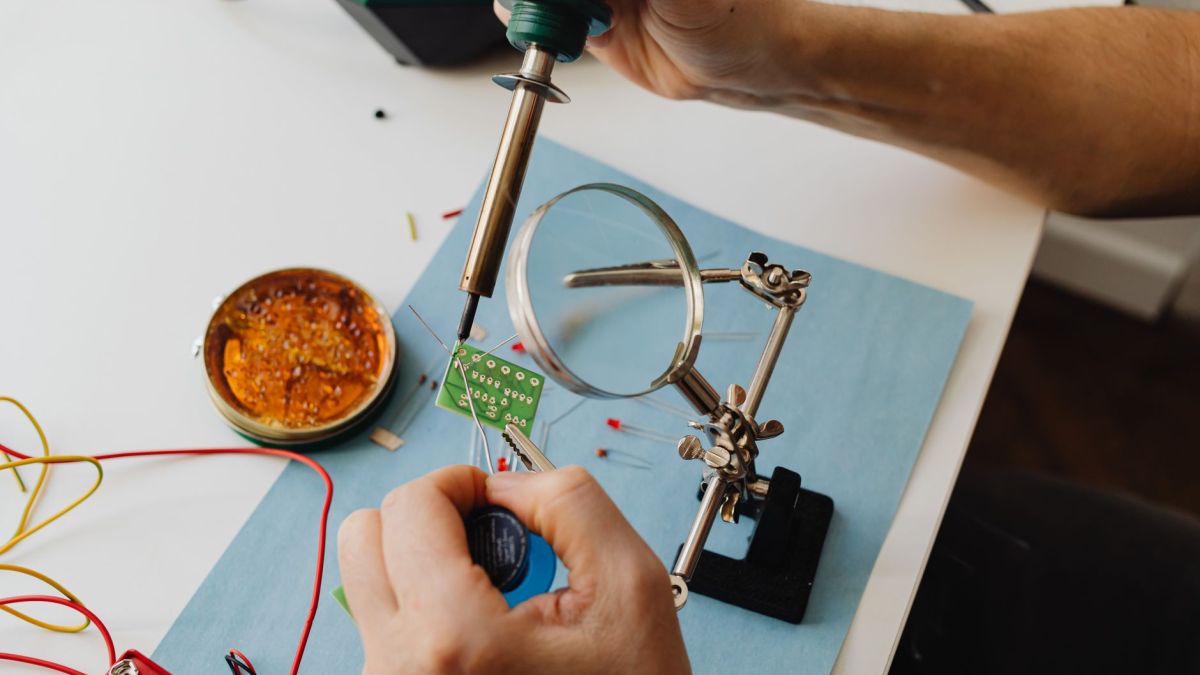 A detailed view of a technician using a soldering iron on a circuit board through a magnifying glass.