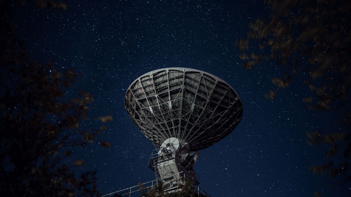 From below of blue starry sky over radio telescope and trees with leaves