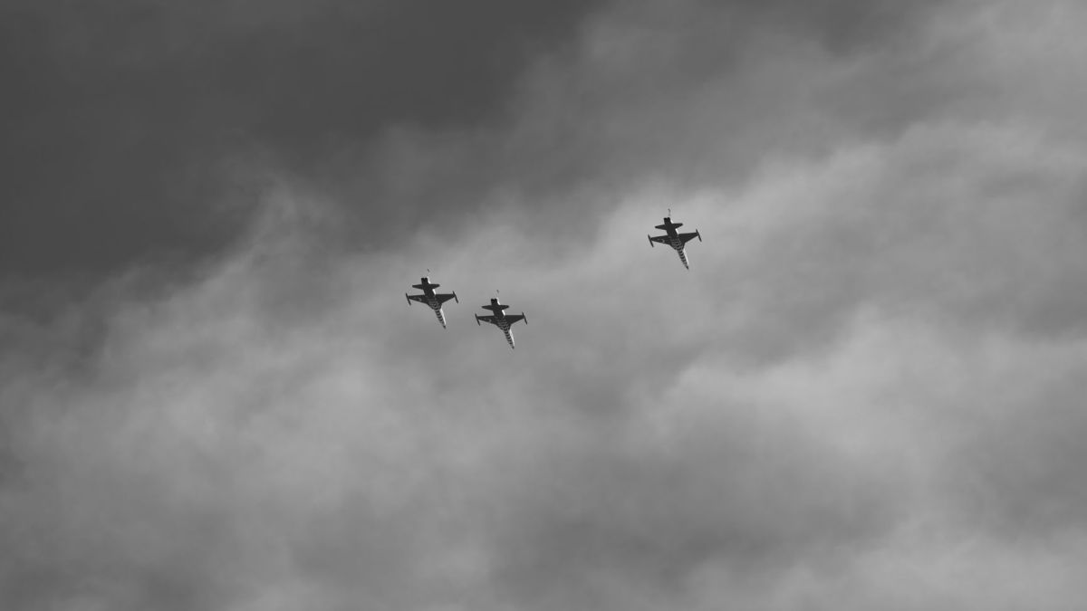 Black and white image of three jets performing aerobatics amidst dramatic clouds.