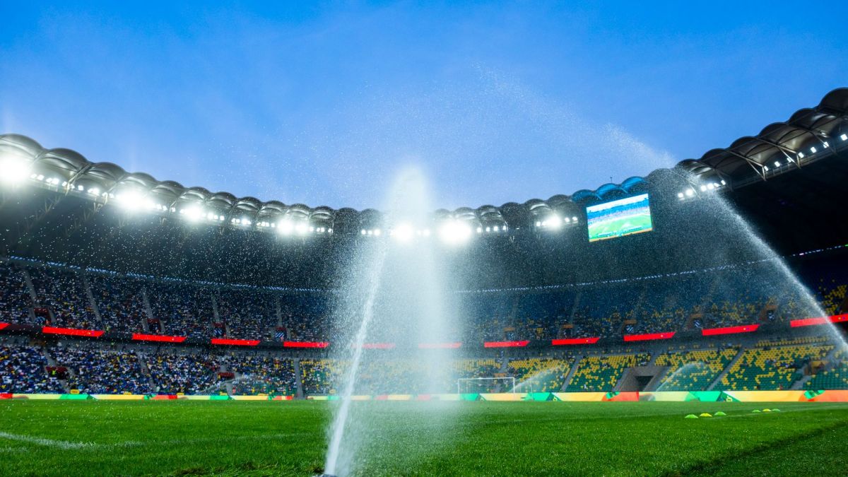 Nighttime view of a soccer stadium with sprinklers watering the field, enhancing the vibrant atmosphere.