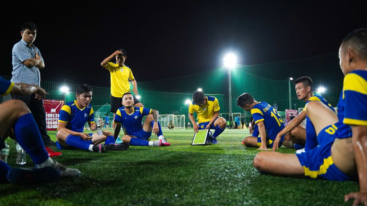 Soccer players in blue and yellow uniforms gather around coach for strategy discussion on field at night.