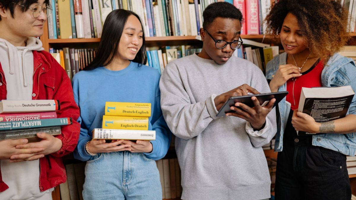 A group of diverse students study together in a library, using books and a tablet for learning.